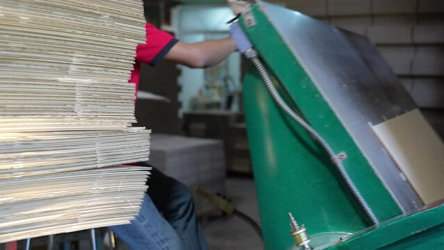 Selective focus of cropped image of Asian male factory worker in a face mask and gloves sitting on a chair putting cardboard paper into a die cutting and creasing machine to make creases on cardboard.