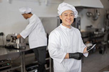 Portrait of smiling asian chef in uniform standing with printed order in professional kitchen with...