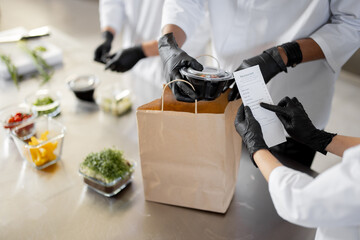 Cooks in protective gloves prepare take away food and packing them into paper bag for delivery, close-up on food. Concept of cooking food at dark kitchen for delivery