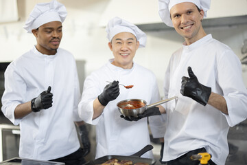 Portrait of three chef cooks with different ethnicities holding pan with a sauce while cooking in the kitchen. Teamwork and cooking delicious food at restaurant. Group of happy diverse cooks together