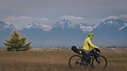 The man travel on mixed terrain cycle touring with bikepacking. The traveler journey with bicycle bags. Sport tourism bikepacking, bike, sportswear in green black colors. Mountain snow capped.