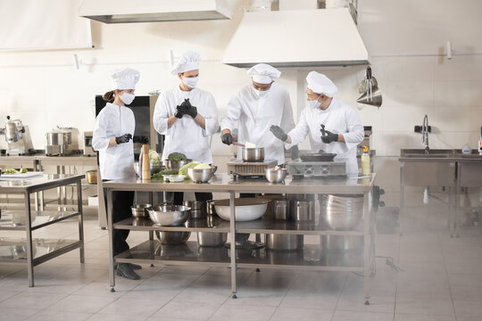 Multiracial team of cooks in white uniform cooking together in restaurant kitchen. Concept of teamwork and hard work at restaurant. Cooks wearing face masks to prevent virus spread  - Powered by Adobe