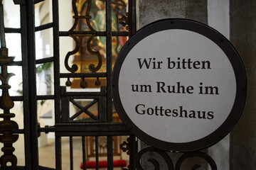 Fototapeta premium A plate with text in German. 'Wir bitten um Ruhe im Gotteshaus' means: We ask for silence in the church. In the background is a blurred church interior.
