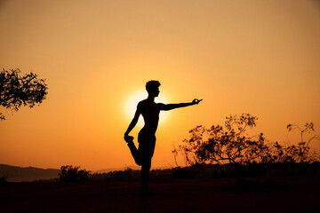 Young man silhouette doing yoga at the sunset on a top of the mountain with the sun behind