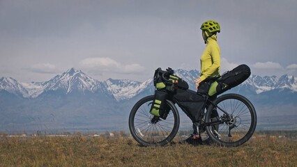 Naklejka premium The woman travel on mixed terrain cycle touring with bikepacking. The traveler journey with bicycle bags. Sport tourism bikepacking, bike, sportswear in green black colors. Mountain snow capped.