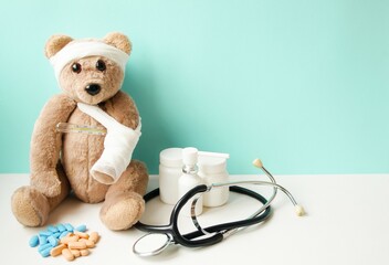 A peach teddy bear with a broken paw and a sore head sits on a turquoise background, next to it is medicine, pills, vitamins and a stotoscope lies.  The concept of children's medicine.  Banner place f