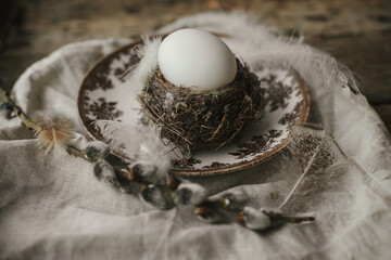 Rustic Easter still life. Natural egg in nest with feathers, vintage plate,  pussy willow branches and napkin on aged wood. Easter table decoration. Moody image