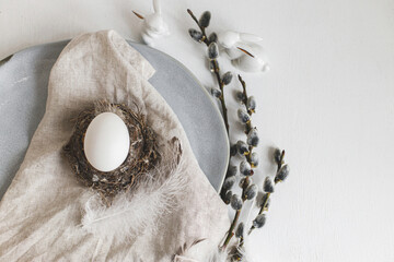 Stylish Easter table setting. Natural easter egg in nest, pussy willow branches, feathers on modern plate with napkin and cutlery on white wooden table. Modern Easter table decoration