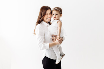 Happy family. Emotional young stylish mother staying and holding in arms laughing adorable toddler baby daughter, having fun playing together, isolated in light room, studio. Happy time of motherhood.