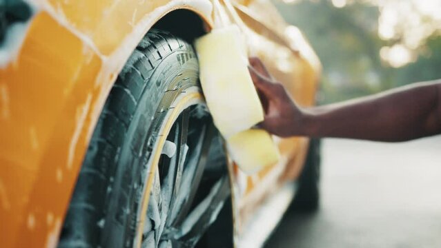 Hands of African man holding yellow sponge, washing car wheel with foam. Cleaning of modern rims of luxury yellow car at self car wash service outdoors. Clean car concept. Foaming after rubbing