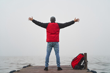 Freedom and success concept. Young man with red backpack opens arms to vast ocean on wooden dock