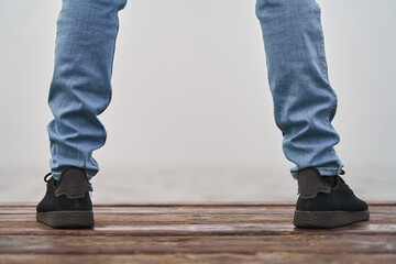 Into the unknown. Detail of legs in jeans and sneakers in front of ocean with fog over wood