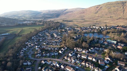 Low level aerial image of a Scottish village and glen surrounded by hills. A bright sunny day with a low sun casting long shadows. Village pond in the middle distance surrounded by houses.