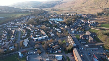 Fototapeta premium Low level aerial image of a Scottish village and glen surrounded by hills. A bright sunny day with a low sun casting long shadows. Village pond in the middle distance surrounded by houses.