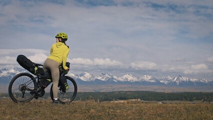 The woman travel on mixed terrain cycle touring with bikepacking. The traveler journey with bicycle bags. Sport tourism bikepacking, bike, sportswear in green black colors. Mountain snow capped.