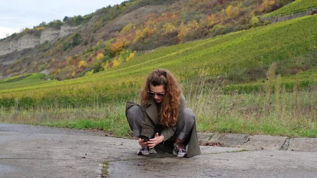Slow motion of a beautiful, stylish hipster woman taking macro photos of insects with her smartphone. Squatting in nature. Autumn landscape, fresh air, freedom and recreation of modern people. 4K