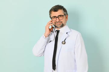 Photo of medical doctor wearing white coat and stethoscope holding mobile phone standing isolated over blue background