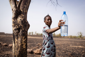 Cute African village girl with a big mouthful of water in her mouth and holding aloft a plastic bottle, recommending the use of clean and fresh drinking water; disease prevention concept - obrazy, fototapety, plakaty