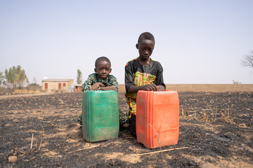 Two sad African boys kneeling in a burnt field behind their large water canisters, staring...