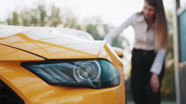 Closeup cropped image of hand of young confident stylish european businesswoman washing and cleaning yellow sport car with microfiber cloth, wiping hood and headlights. Car wash outdoors concept.
