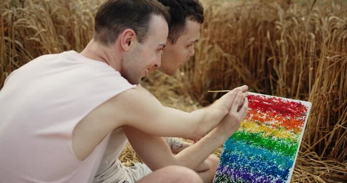 Two Romantic Teenage Gays sitting in the Field and Drawing Rainbow Picture. Using Wheat Stalk Focused on Drawing LGBT Sign. Couple of Men Lovers enjoys Time Together. Nature. Picnic. Artists.