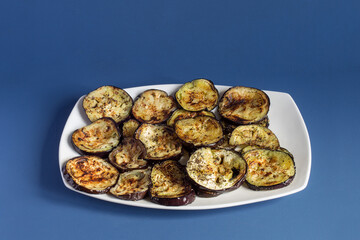 An elegant white platter filled with parsley baked roasted eggplant slices on blue studio background. Presentation and food.