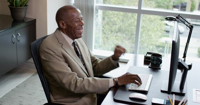 A happy corporate black man types at a desktop computer and laughs to himself near a large office window on a sunny afternoon. 35mm Medium Closeup Tripod Shot 4K.