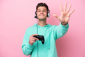 Young handsome caucasian man playing with a video game controller isolated on pink background counting five with fingers