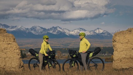 The man and woman travel on mixed terrain cycle bike touring with bikepacking. The two people journey with bicycle bags. Sport sportswear in green black colors. Mountain snow capped, stone arch.