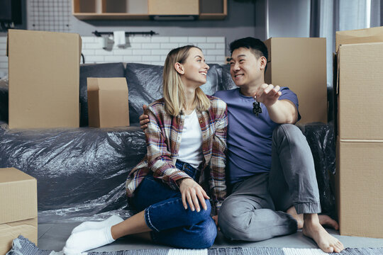 Close-up photo of a young family in a new apartment holding the keys to the house, a multiracial couple sitting in the living room among the cardboard boxes, settled in a new apartment
