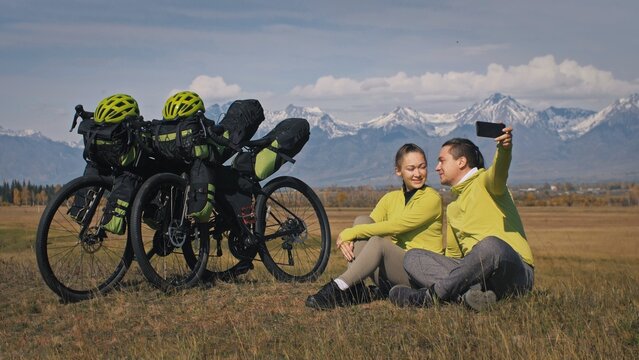 The Man And Woman Travel On Mixed Terrain Cycle Touring With Bikepacking. The Two People Journey With Bicycle Bags. Sport Bikepacking, Bike, Sportswear In Green Black Colors. Mountain Snow Capped.