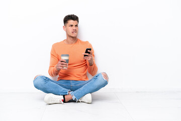 Fototapeta premium Young man sitting on the floor isolated on white background holding coffee to take away and a mobile while thinking something