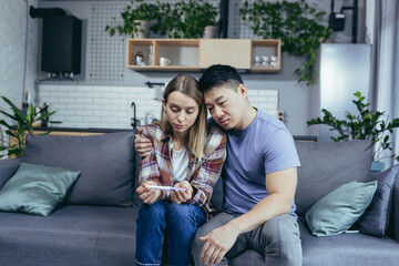 Man Asian supports wife, disappointing pregnancy test result, young multiracial couple sitting on sofa, frustrated