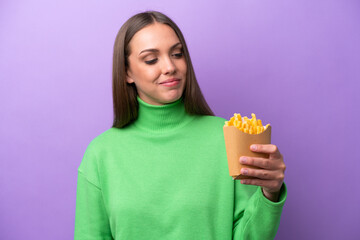 Young caucasian woman holding fried chips on purple background looking to the side and smiling