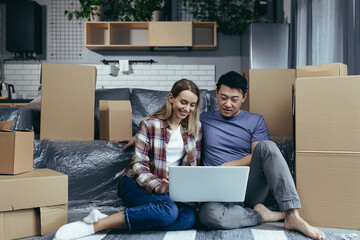 Loving and happy couple sitting on the floor, Asian man and woman among cardboard boxes, looking at...