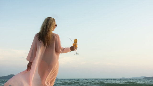 Sexy hot woman walks on golden sunset on tropical beach, wet sand, against background of sea. Girl in green swimsuit and pink tunic silk shirt cape, drinks her orange cocktail Pina Colada.