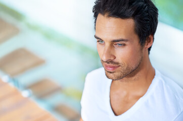 Hes clam and collected. A handsome young man sitting with his back agains a pane of glass.