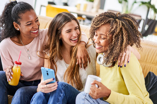 Three cheerful happy woman laughing good using mobile smart phone at home together sitting at sofa drinking juice and coffee. Friends having fun indoors online people - social media