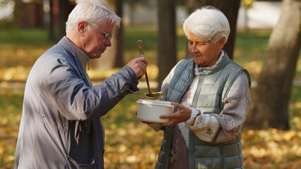 Generous elderly grey-haired caucasian man volunteers in charity organization and pours hot soup...