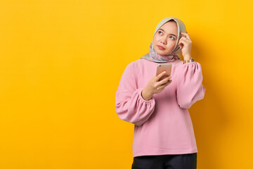 Pensive young Asian woman in pink shirt using smartphone thinking about a question on yellow background