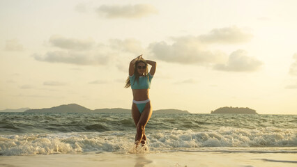 Sexy woman walking alone on beach coastline and relax warm golden sunset. Woman walking on water in blue swimsuit and sunglasses. Concept rest tropical resort traveling tourism happy summer holidays