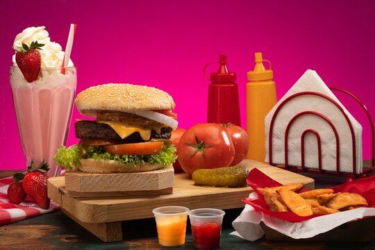Hamburguesa, condimentos y malteada en fondo rosa acompa&ntilde;ado con props