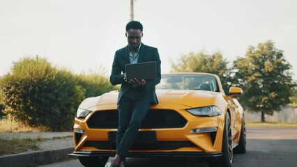 Handsome African American man in a business suit. Businessman working with laptop. Man standing near his yellow sport car on the background of green park