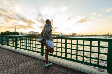 Fototapeta premium Confidence African man in sportswear stretching his body while jogging exercise on the bridge in the city at summer sunset. Strong male athlete enjoy outdoor activity sport training running workout