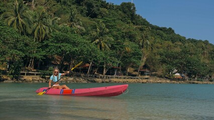 Fototapeta na wymiar Young blonde woman in blue swimsuit rows pink plastic canoe along azure sea bay past island with palms under blue sky at resort. Traveling to tropical countries. Girl is sailing on kayak in ocean.