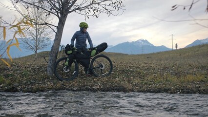 The woman travel on mixed terrain cycle touring with bikepacking. The traveler journey with bicycle bags. Sportswear in green black colors. The trip in magical autumn forest, river stream.