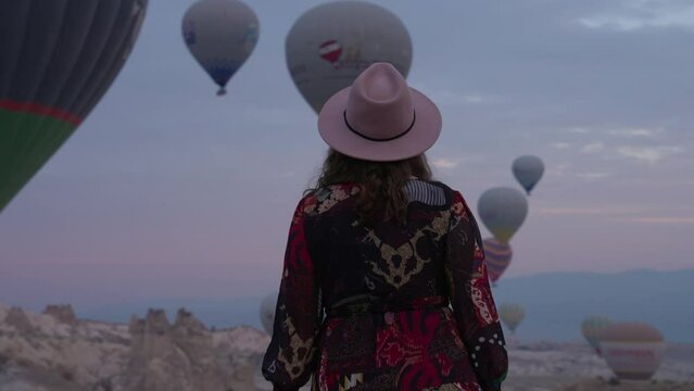 Fashionable Girl In Dress And Hat Watching The Sky At Sunrise Full Of Colorful Hot Air Balloons In Cappadocia, Turkey. - medium shot