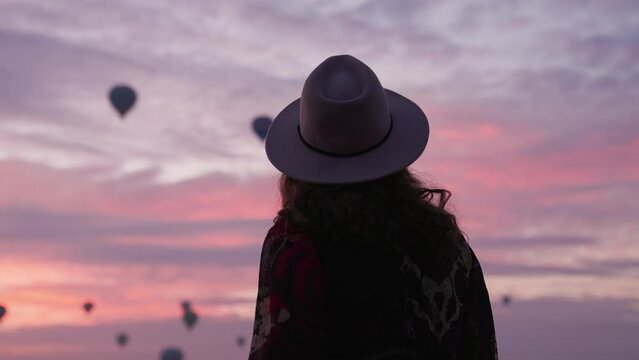 Young Girl Admiring The Hot Air Balloons Flying In Cappadocia At Sunrise In Turkey. - rear view