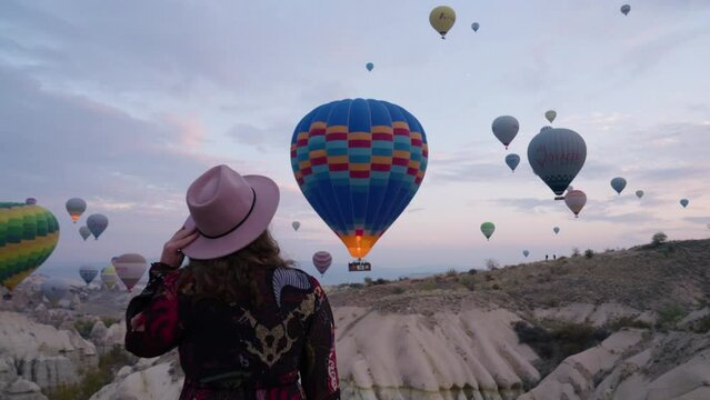 Female Tourist In Long Dress And Hat Watching Colorful Hot Air Balloons Flying In The Sky At Sunrise In Cappadocia, Turkey. Pullback