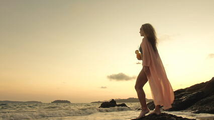 Fototapeta na wymiar Silhouette woman stand on a reef rock stone in sea on golden sunset. Girl on tropical beach in green swimsuit and flutter in wind pink tunic silk shirt cape, drinks her orange cocktail Pina Colada